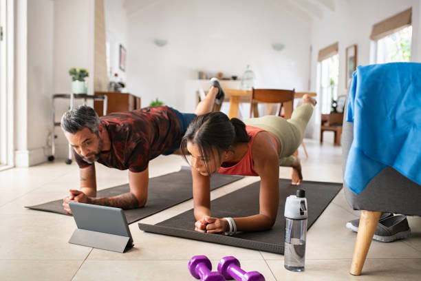 Multiethnic mature couple exercising at home and watching training videos on digital tablet. Middle aged woman and indian man doing planks with a leg outstretched while watching fitness lessons online on digital tablet. Fit mid adult couple doing strenght plank following online tutorials. , personal training , chennai , busy people , personal training