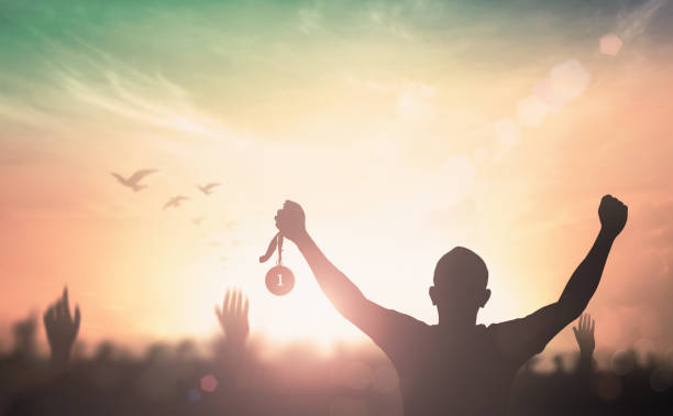Silhouette champion hand holding gold medal reward against blurred sport crowd autumn sunset background