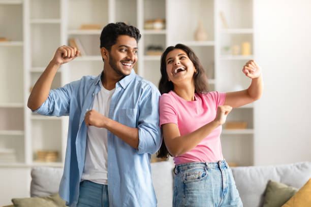 Joyful hindu spouses dancing in living room interior at home, happy indian couple having fun and laughing, husband and wife enjoying spending time together